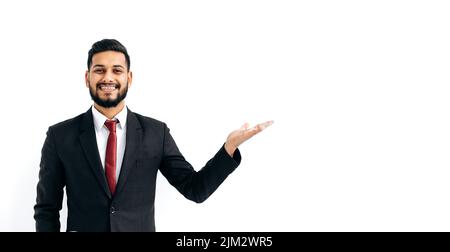 Homme arabe ou indien élégant positif en costume formel, debout sur un arrière-plan blanc isolé, regardant la caméra, pointant la main vers l'espace vide, souriant joyeusement. Espace de copie pour votre présentation Banque D'Images