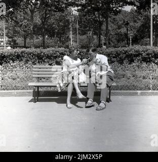 1950s, historique, un jour ensoleillé et une dame assise sur un banc à côté d'un homme, éclairant sa cigarette, en utilisant sa propre cigarette allumée, Angleterre, Royaume-Uni. Banque D'Images