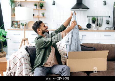 Un homme indien ou arabe souriant se dévêtue de son paquet tant attendu, examine les vêtements, heureux d'un bon achat, se trouve à la maison sur le canapé dans le salon. Concept d'achat en ligne Banque D'Images