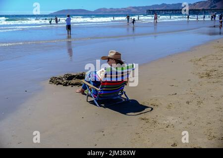 Lady in Sun Hat est assis dans une chaise au bord de l'eau en lisant un livre à Pismo Beach, Californie, avec des montagnes et Pismo Pier en arrière-plan. Banque D'Images