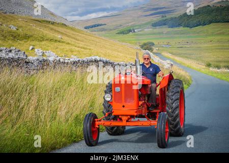 31.07.2022 Ingleton, North Yorkshire, UK Un homme portant des lunettes de soleil, une chemise bleue conduisant un tracteur rouge vintage dans les Yorkshire Dales Banque D'Images