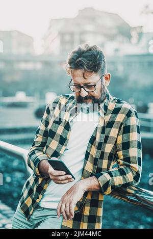 Homme mature écrit et lit sur un téléphone mobile debout à l'extérieur. La technologie et les gens modernes concept de style de vie. Style taille basse avec un homme à barbe et des lunettes Banque D'Images