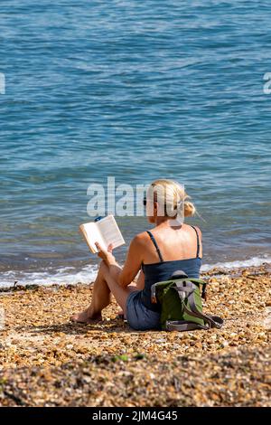 jeune femme attrayante portant une robe d'été lisant un livre assis sur une plage de galets pendant une vague de chaleur d'été, femme assise au soleil sur la plage. Banque D'Images