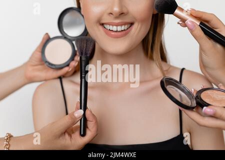 Gros plan photo d'une fille souriante heureuse avec maquillage parfait et mise en valeur des pommettes sur fond blanc. Maquilleuses appliquant du maquillage avec des pinceaux Banque D'Images