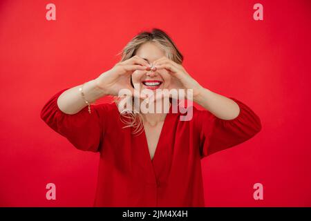 Portrait d'une femme heureuse avec un sourire à la neige blanche en rouge de vacances avec un maquillage lumineux parfait isolé sur fond rouge. Expression de l'amour, faire le coeur Banque D'Images