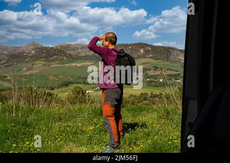 man with backpack observing the horizon mountain landscape in the background and cloudy sky seen from inside his van Stock Photo