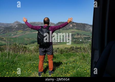 man on his back with backpack with arms outstretched mountain in the background clear blue sky ,seen from inside his van Stock Photo