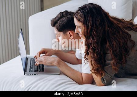 Couple amoureux se trouve sur le canapé derrière un ordinateur portable dans le salon près de la fenêtre et fait des achats en ligne ou prévoit un voyage Banque D'Images