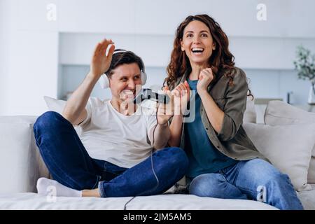Une jeune femme excitée soutient et embrasse son mari dans un casque jouant à des jeux en ligne avec télécommande dans le salon Banque D'Images