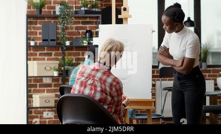 Un jeune artiste donne une leçon de dessin à un élève aîné de l'école d'art, expliquant comment dessiner un modèle de vase sur toile. Assister à un atelier créatif pour dessiner des œuvres d'art avec des instruments artistiques. Banque D'Images