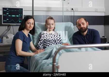 Portrait de parents soignés assis à côté d'une fille malade hospitalisée se reposant dans le lit du patient de l'établissement de santé des enfants tout en regardant l'appareil photo. Famille caucasienne à l'intérieur de la salle pédiatrique de l'hôpital. Banque D'Images