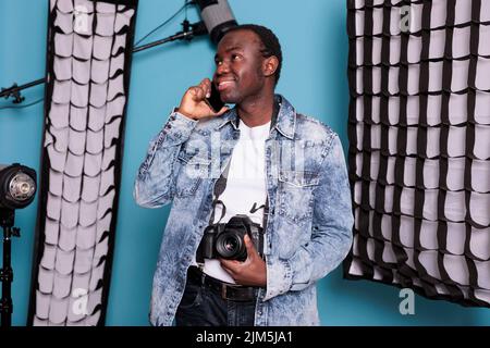 Photographe professionnel parlant sur smartphone en studio avec des softbox et des équipements de production. Producteur vidéo créatif sur fond bleu avec projecteur pendant un appel téléphonique. Banque D'Images