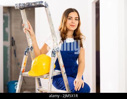 Jeune femme enthousiaste bâtisseur en combinaison bleue et chapeau de maçon jaune Banque D'Images