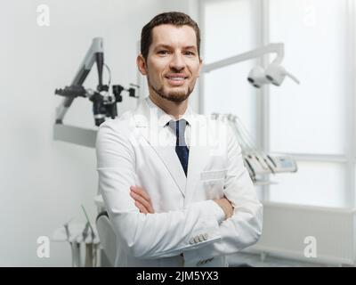 Joyeux dentiste de sexe masculin en blouse de laboratoire blanche posant avec les mains croisées et regardant la caméra Banque D'Images