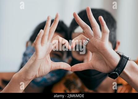 Par la maladie et la santé. Un jeune couple masqué faisant un geste en forme de coeur avec leurs mains à la maison. Banque D'Images