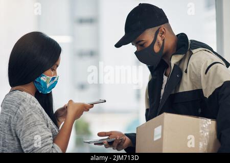 Le paiement sans contact permet de garder le contrôle. Un jeune homme et une jeune femme masqués utilisant un smartphone lors d'une livraison à domicile. Banque D'Images