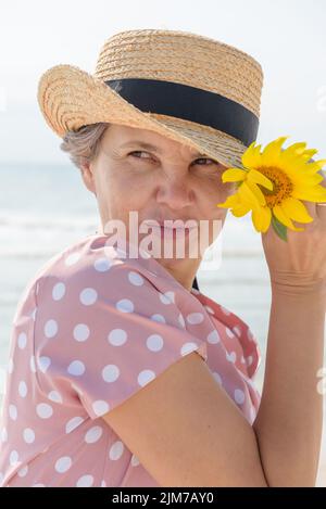 Femme dans une robe rose avec des pois blancs tenant chapeau de paille et tournesol dans sa main, regardant loin et cutly souriant, sur fond de mer. Banque D'Images