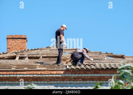 Région de Krasnodar. Russie. 15 juillet 2022. Les couvreurs mâles en uniforme retirent les anciennes feuilles d'ardoise de toiture du toit d'un bâtiment. Les travailleurs réparent le toit d'un bâtiment par temps ensoleillé. Banque D'Images