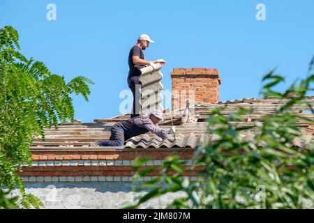 Région de Krasnodar. Russie. 15 juillet 2022. Les couvreurs mâles en uniforme travaillent sur le toit d'un bâtiment. Les travailleurs démonte les anciennes feuilles d'ardoise d'amiante. Banque D'Images