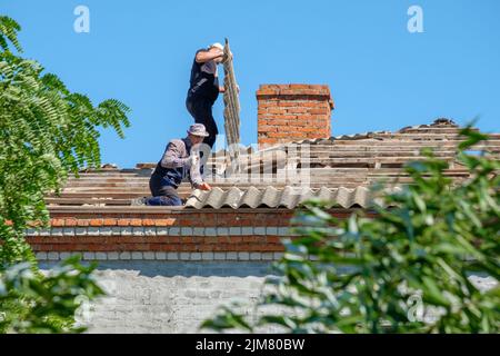 Région de Krasnodar. Russie. 15 juillet 2022. Les ouvriers de toiture en uniforme réparent le toit d'un bâtiment par un jour ensoleillé. Les hommes retirent les anciennes feuilles d'ardoise de toiture du toit d'un bâtiment. Banque D'Images