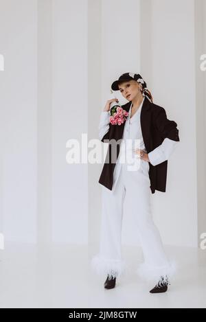 Une jeune femme élégante avec une tenue tendance, un costume blanc clair avec des plumes, une veste noire, une casquette de baseball et un bouquet de fleurs entre ses mains Banque D'Images