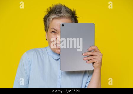 En ligne troll femme mature se cache derrière la tablette numérique avec l'expression faciale maléfique isolée sur fond jaune. Femme moderne senior poster de mauvais commentaires en ligne et heureux à son sujet. Haine, concept intimidateur. Banque D'Images
