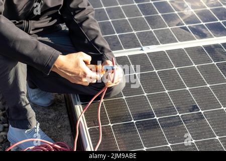 Vue rapprochée d'un homme de travail méconnaissable, les mains coupant un fil électrique rouge avec une pince et des entonnoirs solaires en arrière-plan Banque D'Images