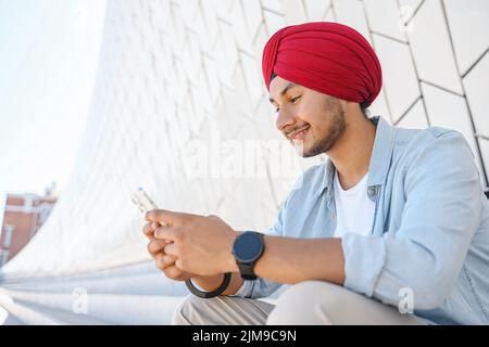 Portrait de jeune Indien gai en turban envoyant un message sur téléphone portable assis sur les marches à l'extérieur. Heureux hindou utilisant un smartphone, en appréciant le chat en ligne, la messagerie sur les réseaux sociaux Banque D'Images