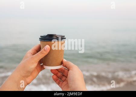 Café à la mer. Une jeune femme tient une tasse de café jetable sur un matin ensoleillé à la plage. Banque D'Images