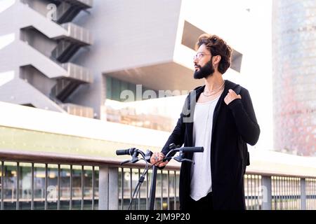 jeune homme caucasien avec barbe portant des vêtements tendance et marchant avec son vélo pliant à travers la ville, concept de style de vie urbain et durable m. Banque D'Images