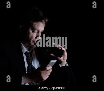 Homme mûr stressé buvant une bière tout en vérifiant son téléphone cellulaire dans l'obscurité Banque D'Images
