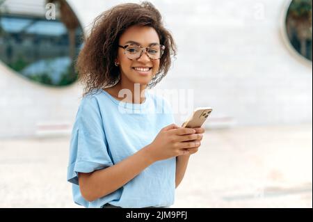 Bonne charmante jeune femme afro-américaine avec des lunettes, portant une chemise bleue, se tient à l'extérieur près du centre d'affaires, utilise son smartphone, envoie des SMS avec des amis ou des collègues, regarde l'appareil photo, sourit Banque D'Images