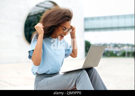 Jeune femme afro-américaine pleine d'émotions et de gaieté avec des lunettes, utilise un ordinateur portable tout en étant assise à l'extérieur, se fait des mains gestives, regarde l'écran avec enthousiasme, lit des nouvelles inattendues, célèbre le succès, sourit Banque D'Images