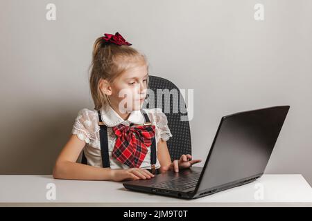 Une petite fille dans un uniforme d'école utilise un ordinateur portable à une table sur un fond blanc, l'espace de copie Banque D'Images