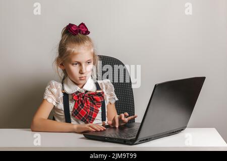 Une petite fille dans un uniforme d'école utilise un ordinateur portable à une table sur un fond blanc, l'espace de copie Banque D'Images