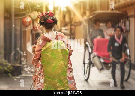 Arrière d'une femme Maiko marchant sur un chemin de la ville de Kyoto, au contre-jour de la lumière du coucher du soleil traversant un pousse-pousse. Banque D'Images