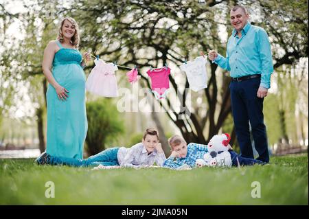 Happy pregnant couple avec deux fils holding girls' vêtements de bébé sur fond de chaîne nature printemps Banque D'Images