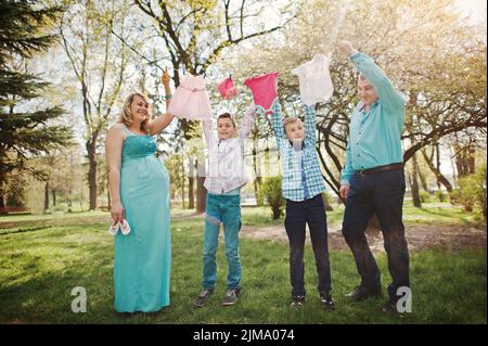 Happy pregnant couple avec deux fils holding girls' vêtements de bébé sur fond de chaîne nature printemps Banque D'Images