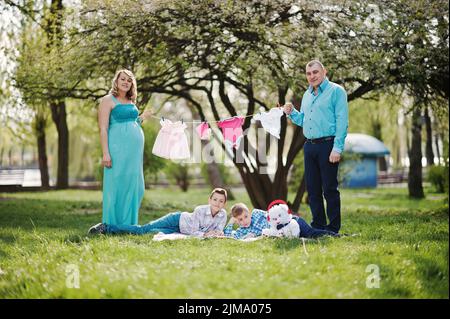 Happy pregnant couple avec deux fils holding girls' vêtements de bébé sur fond de chaîne nature printemps Banque D'Images