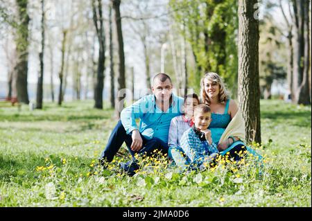 Happy pregnant famille avec deux fils, vêtu d'un vêtements turquoise assis sur l'herbe avec des fleurs au park Banque D'Images