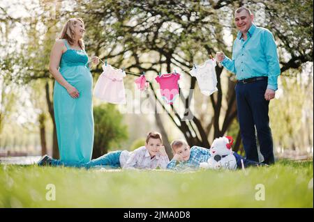 Happy pregnant couple avec deux fils holding girls' vêtements de bébé sur fond de chaîne nature printemps Banque D'Images