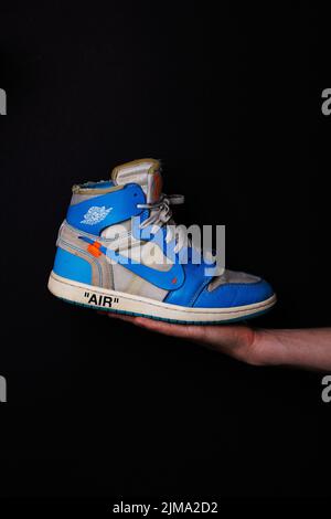 Main d'un homme tenant la sneaker Off-White Air Jordan 1 Retro High OG 'UNC' sur fond noir Banque D'Images