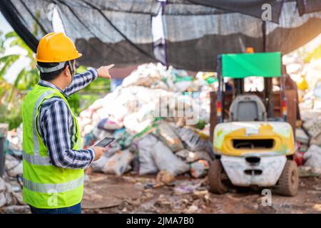 Contremaître asiatique travaillant dans une usine de recyclage et tenant des tablettes et regardant l'usine de recyclage des déchets avec un chariot élévateur à fourche sur le fond. Banque D'Images