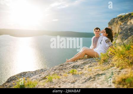 Couple portant sur l'habillement avec un modèle assis au beau paysage, sur la falaise au coucher du soleil dans l'amour. Banque D'Images