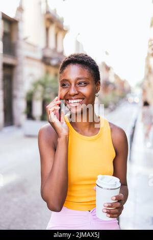 Femme africaine positive discutant par téléphone dans la rue. Elle a des cheveux très courts et élégants, des vêtements décontractés d'été et tient une tasse de café réutilisable Banque D'Images