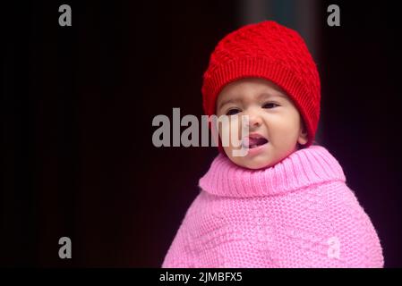 Une adorable petite fille indienne de 13 mois portant une casquette d'hiver rouge et un poncho Banque D'Images