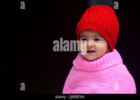 Une adorable petite fille indienne de 13 mois portant une casquette d'hiver rouge et un poncho Banque D'Images