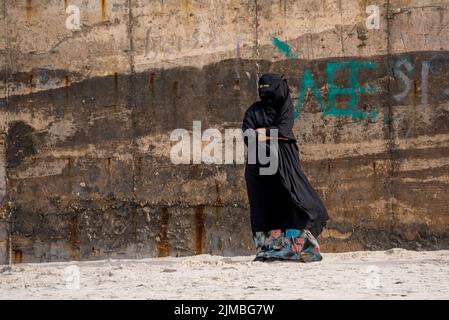 La femme dans une abaya traditionnelle noire avec un voile debout devant un mur grungy. Banque D'Images