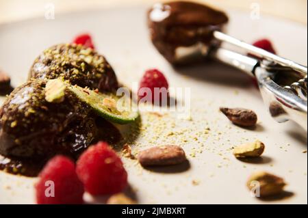 Plat de boules de glace au chocolat surgelées, garnies de pistaches, de fèves de cacao, de framboises et d'une tranche de lime juteuse. Boule en métal effet flou bourrée d'humour Banque D'Images