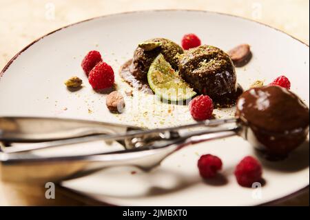 Accent sélectif sur les boules de glace au chocolat surgelées sur une assiette blanche garnie d'une tranche de lime juteuse, de pistaches, de framboises et de fèves de cacao an Banque D'Images